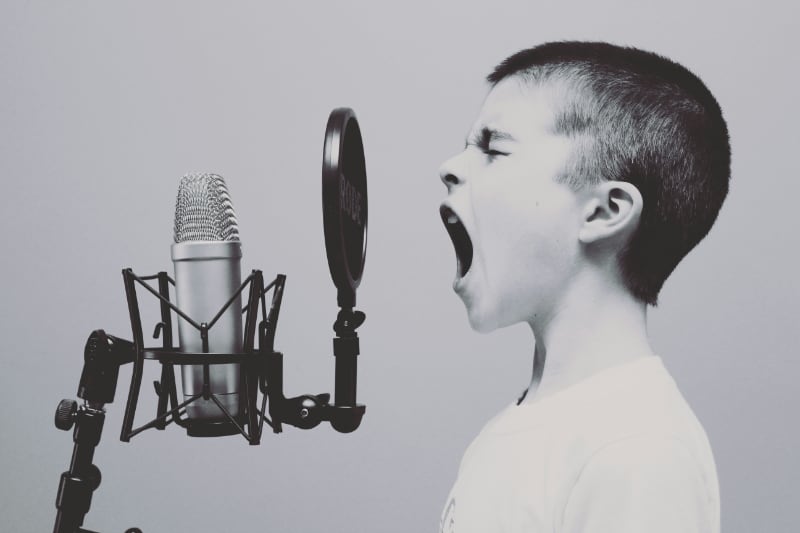  A black and white foto of a young kid singing into a microphone. You see the kid from the side standing in front of a pop shield. 