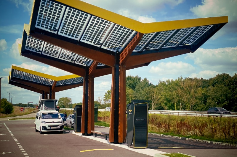  A white van parked at a electric charing station. The station has a solar covered roof. The van has opened it's pop-up roof to make it possible to nap while charging 